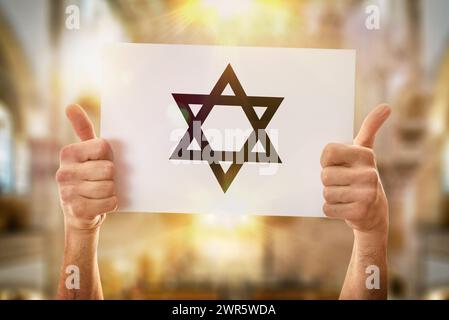 Mani di un uomo religioso con un buon cartello che regge un poster bianco con una stella ebrea disegnata con sfondo sinagoga Foto Stock