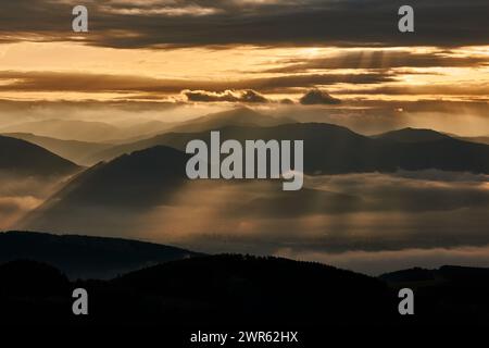 Paesaggio nebbioso di montagna all'alba. I primi raggi del sole brillano attraverso le nuvole colorate. Con una silhouette di colline. Vrsatec, Slovacchia Foto Stock