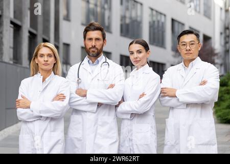 Ritratto di un gruppo interrazziale di medici e scienziati, uomini e donne in camice medico bianco in piedi fuori da una clinica, attraversando le braccia sopra il petto e guardando seriamente la telecamera. Foto Stock