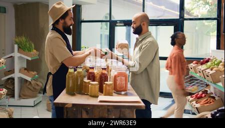 Shopper presenta condimenti e prodotti in vasetti di vetro ai clienti vegani mentre acquistano generi alimentari. L'uomo del Medio Oriente vuole comprare frutta e verdura fresca biologica. Foto Stock