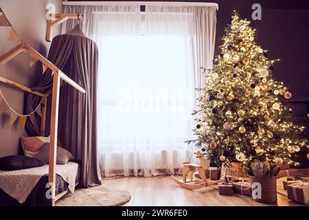 Decorazioni natalizie in un accogliente soggiorno con albero di natale con corde luminose, sedia a dondolo in legno, regali, letto con baldacchino, bandiere per feste. Sfondo con spazio di copia. Foto Stock