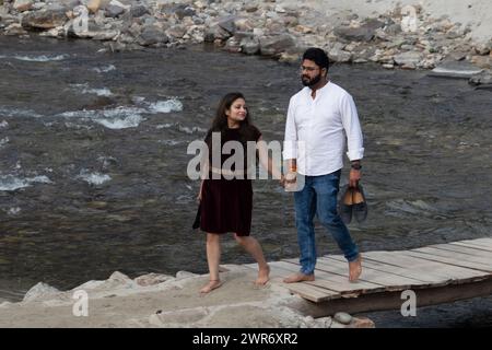 Una coppia passeggia mano nella mano su una passerella sul fiume Gange a Rishikesh, trasportando le loro calzature attraversano con grazia il ponte insieme Foto Stock