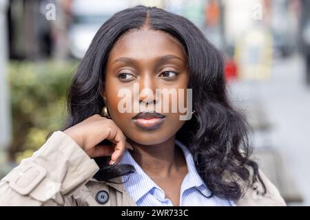 La fotografia mostra una giovane donna africana in un pensiero contemplativo in mezzo a uno sfondo urbano. È vestita in modo intelligente con un trench su una camicia a righe blu, che incarna un mix di moda professionale e casual. La sua mano tocca delicatamente il viso, suggerendo introspezione o un momento di pausa nella sua giornata intensa. I suoi capelli ondulati e il suo delicato trucco mettono in risalto la sua bellezza naturale, mentre guarda leggermente fuori dalla fotocamera, indicando un pensiero profondo o un coinvolgimento con l'ambiente circostante. Giovane donna riflessiva in città all'aperto. Foto di alta qualità Foto Stock