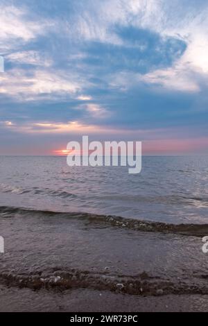 Questa fotografia cattura la tranquilla maestosità di un tramonto sul mare, con il sole che si tuffa sotto l'orizzonte, la sua luce diffusa da un velo di nuvole. Il cielo è una tela di blues tenui e rosa tenui, che riflette l'ultimo saluto del sole alla giornata. Il mare, dolce e soffocato, lambisce la costa rocciosa, contribuendo a creare un'atmosfera serena. Le nuvole sono striate artisticamente attraverso il cielo, aggiungendo profondità e consistenza alla visualizzazione celeste. Questa scena evoca un senso di calma e introspezione mentre il giorno passa alla notte. Deck's Delight: Tramonto tranquillo sul mare dolce. Foto di alta qualità Foto Stock