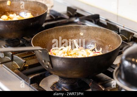 Questa immagine dà un'occhiata dietro le quinte di una cucina commerciale con due fornelli a gas, uno con cipolle metà saltata e l'altro con funghi. I woks appaiono ben stagionati e utilizzati, come è tipico di una cucina professionale dove velocità ed efficienza sono fondamentali. Lo sfondo della cucina, anche se fuori fuoco, trasmette un ambiente di cucina molto affollato. Commercial Kitchen Scene with Woks on Stoves. Foto di alta qualità Foto Stock