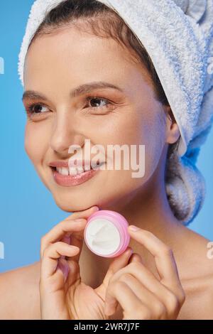 donna gioiosa con pelle lucente e asciugamano che regge il vaso di crema cosmetica e guarda lontano il blu Foto Stock