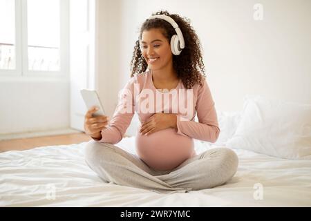 Donna incinta nera felice che indossa le cuffie utilizzando lo smartphone a casa Foto Stock