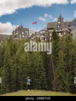 Mia moglie si trova in primo piano su alcuni alti pini e il castello nel cielo noto anche come Fairmont Banff Springs. Foto Stock