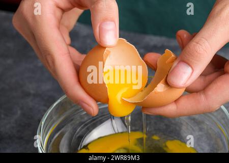 La casalinga ha le mani spaccando uova fresche, tuorli e bianchi in una ciotola. La mano di una donna che spacca un uovo con un coltello in una ciotola di vetro trasparente nella k grigia Foto Stock