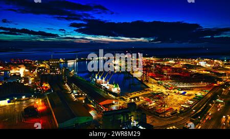 Vista notturna panoramica di un vivace porto industriale con gru, container ed edifici illuminati contro un cielo crepuscolo a Liverpool, Regno Unito. Foto Stock