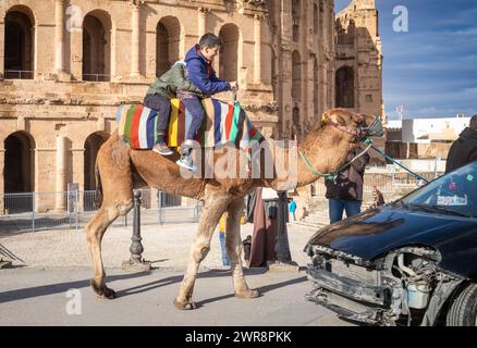 Due giovani ragazzi fanno un giro in cammello all'esterno dell'anfiteatro romano El Jem (Thysdrus), in Tunisia. Foto Stock