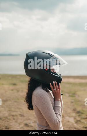 Profilo laterale di una donna che regola il suo casco integrale da moto, con un lago sereno e un cielo nuvoloso sullo sfondo Foto Stock
