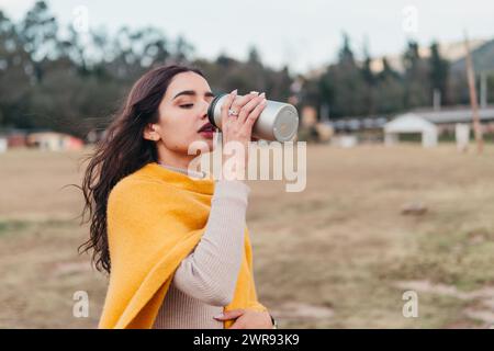 Una giovane latina sorseggiando una tazza da viaggio, avvolta da un maglione giallo accogliente, all'aperto con la natura sullo sfondo Foto Stock