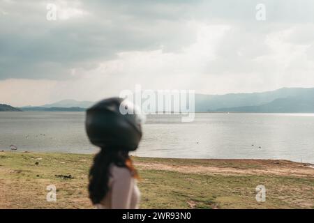Vista laterale di una donna che indossa un casco da moto, che guarda un lago e una catena montuosa tranquilli, incarnando un senso di avventura. Foto Stock