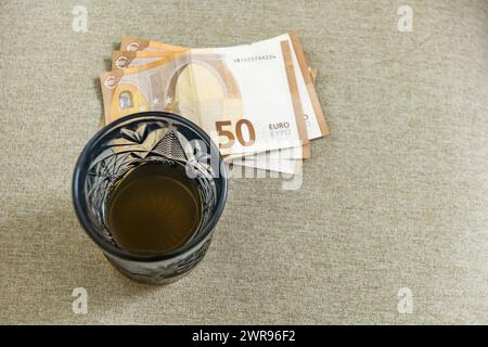 Un vetro di cristallo blu tagliato con liquido ambrato all'interno accanto a un pezzetto di banconote euro su un sedile rivestito in tessuto beige Foto Stock