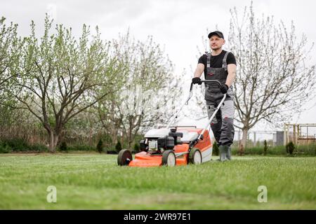 Il giardiniere professionista in abbigliamento protettivo taglia prati verdi utilizzando un moderno rasaerba a benzina senza fili nel cortile posteriore. Giardino stagionale desi Foto Stock