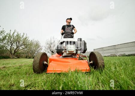 Il giardiniere professionista in abbigliamento protettivo taglia prati verdi utilizzando un moderno rasaerba a benzina senza fili nel cortile posteriore. Giardino stagionale desi Foto Stock