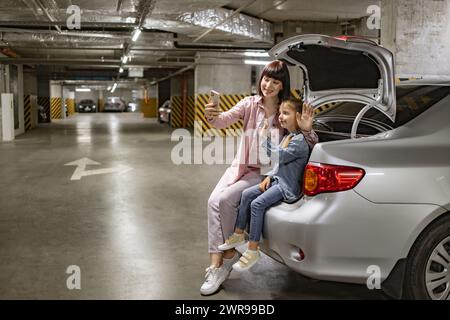 La famiglia indossa abiti casual che effettuano videochiamate utilizzando il cellulare seduto sul bagagliaio Foto Stock