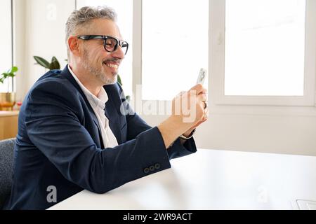 Vista laterale di un dipendente uomo d'affari sorridente e maturo che utilizza uno smartphone moderno, messaggi di testo, messaggistica tramite connessione Internet wireless, navigazione su smartphone senior in un ufficio moderno Foto Stock