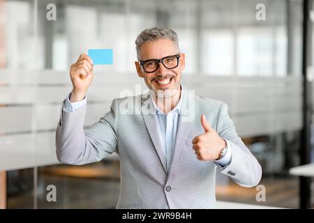 Un uomo d'affari maturo ed elegante regge un biglietto da visita vuoto, il che significa che è pronto per la rete e gli scambi professionali. Il suo gesto di pollice in su indica approvazione e un approccio aziendale positivo. Foto Stock