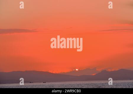 Cielo cremisi e sole a sfera al tramonto sul mare dietro le colline Foto Stock