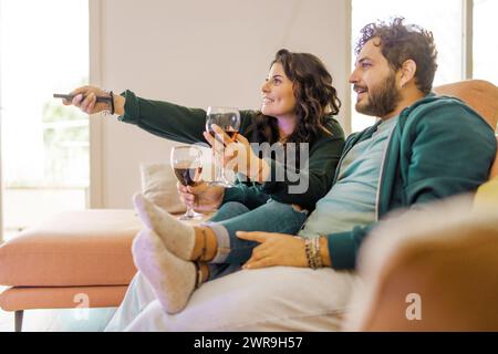 Una coppia sorridente gode di uno spettacolo televisivo insieme durante una confortevole serata a casa, ognuno con un bicchiere di vino rosso - una foto di blis domestico Foto Stock