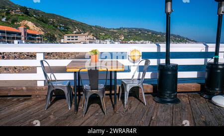 Tavolo e sedie sul molo vicino all'oceano. Tavolo vuoto per due con vista sulla spiaggia, molo costiero con collina e montagne sullo sfondo a Malibu Foto Stock