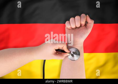 La testa di uno stetoscopio medico sul polso (primo piano) sullo sfondo della bandiera tedesca. Concetto di sistema sanitario tedesco Foto Stock