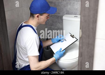 Giovane idraulico che scrive i risultati dell'esame della ciotola del water nell'armadio ad acqua Foto Stock