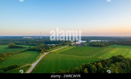 Questa immagine aerea cattura la quiete dell'alba che si infrangono su un vasto paesaggio dedicato alle energie rinnovabili. In primo piano, una strada asfaltata attraversa vivaci campi verdi, che conducono verso turbine eoliche lontane che si stagliano contro il cielo che si risveglia. La fusione di elementi naturali e ingegnerizzati parla di una coesistenza sostenibile. Il delicato gradiente dal cielo caldo alla terra più fresca suggerisce il rinvigorente inizio di una nuova giornata. L'alba sul panorama delle energie rinnovabili. Foto di alta qualità Foto Stock