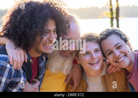 Questa coinvolgente immagine cattura un gruppo di amici che condividono una risata insieme. La composizione include un mix diversificato di individui, tra cui un giovane con i capelli ricci, un uomo più anziano con la barba bianca e gli occhiali, e due donne, che si godono la compagnia reciproca. La delicata attenzione sullo sfondo con il lago e la luce del sole suggerisce un ambiente all'aperto rilassato, enfatizzando ulteriormente lo spirito gioioso e spensierato del gruppo. Gruppo Close-Knit che condivide le Risate in un ambiente illuminato dal sole. Foto di alta qualità Foto Stock