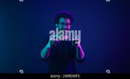 Ritratto in neon ultravioletto felice indiano ragazzo arabo uomo operaio codificatore hacker ingegnere informatico fare gesto hey si mostra dito indice davanti Foto Stock