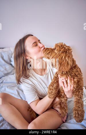 una giovane donna che gioca e bacia una ragazza cockapoo sul letto, minimalismo Foto Stock