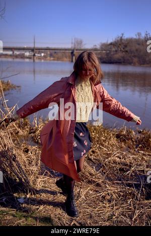 la giovane donna elegante in trench rosa balla sulla riva di un fiume primaverile Foto Stock