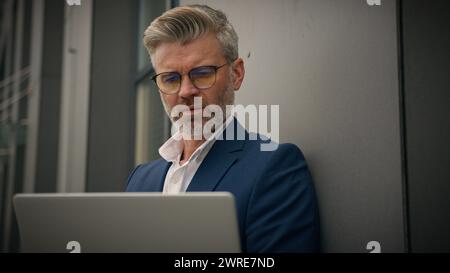 Caucasico anziano senior man maturo business manager occupato datore di lavoro di mezza età uomo d'affari pendente muro edificio per uffici scrivendo computer portatile funzionante Foto Stock