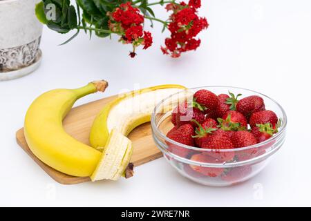Banane di frutta e fragole in una ciotola di vetro con fiore rosso sullo sfondo, concetto di consumo di frutta fresca Foto Stock