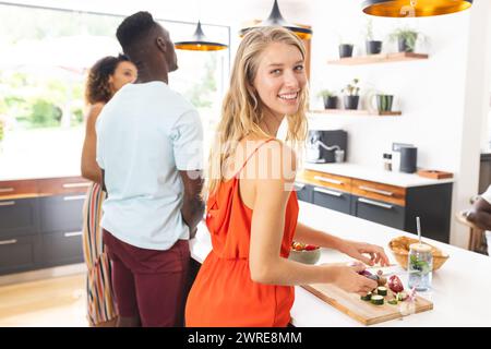 La giovane donna caucasica con un vestito arancione prepara il cibo, sorridendo alla macchina fotografica Foto Stock