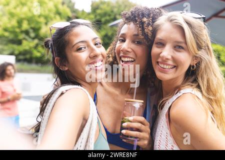 Tre diverse amiche sorridenti si fanno un selfie, una che tiene un drink Foto Stock