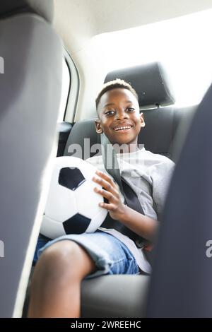 Un ragazzo afroamericano siede in un'auto con un pallone da calcio, sorridendo alla telecamera Foto Stock