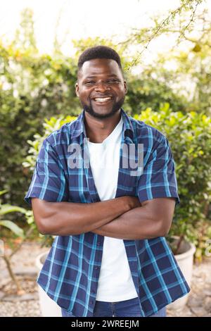 L'uomo afroamericano sorride in grande, indossa una camicia blu a quadri e sta in piedi all'aperto in un giardino Foto Stock