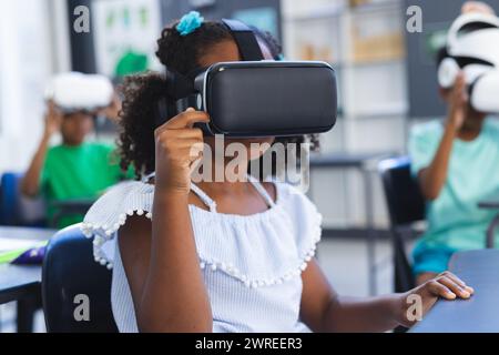 Ragazza birazziale con capelli ricci esplora la realtà virtuale a scuola, compagni di classe in background Foto Stock