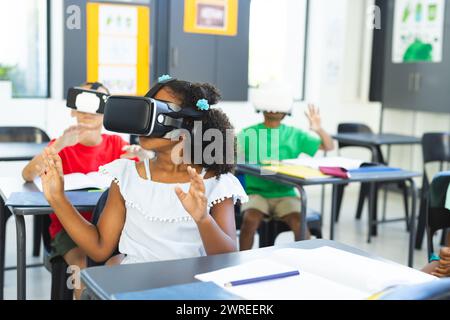 Una ragazza birazziale con visore VR alza la mano in una classe a scuola, circondata da colleghi Foto Stock