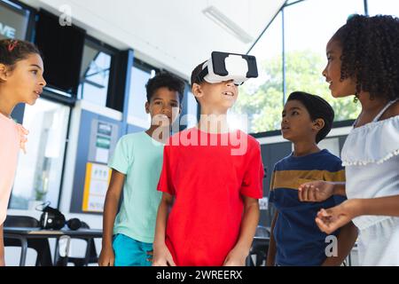 I bambini birazziali sono riuniti intorno a un ragazzo utilizzando un visore per la realtà virtuale in una classe a scuola Foto Stock