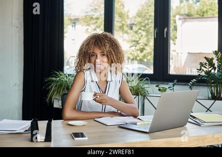 felice donna d'affari afroamericana con apparecchi che tengono una tazza di caffè accanto al computer portatile in ufficio Foto Stock