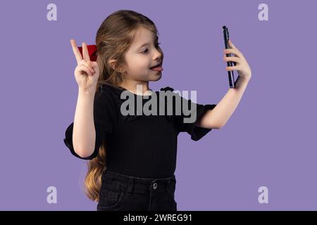 Foto di una graziosa e affascinante bambina con t-shirt nera che scatta selfie con un dispositivo moderno con sfondo viola isolato con il segno V. Alta qualità fot Foto Stock