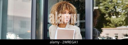 felice donna d'affari afroamericana con capelli ricci che tengono la cartella e escono dall'edificio, striscione Foto Stock
