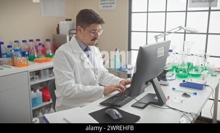 Scienziato ispanico che lavora al computer in un moderno ambiente di laboratorio con becher e provette Foto Stock