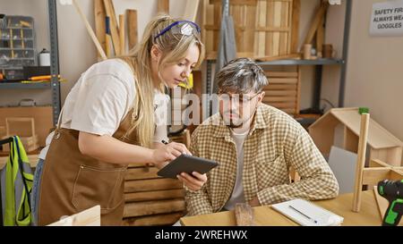 Un uomo e una donna esaminano insieme un tablet digitale in un laboratorio di falegnameria ben attrezzato, mostrando lavoro di squadra e artigianato. Foto Stock