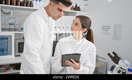 Uomo e donna in camici da laboratorio che discutono su un tablet digitale in un laboratorio scientifico pieno di attrezzature. Foto Stock