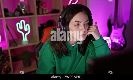 Giovane donna concentrata che gioca in interni di notte, con luci al neon che le illuminano il viso sereno e cuffie moderne. Foto Stock